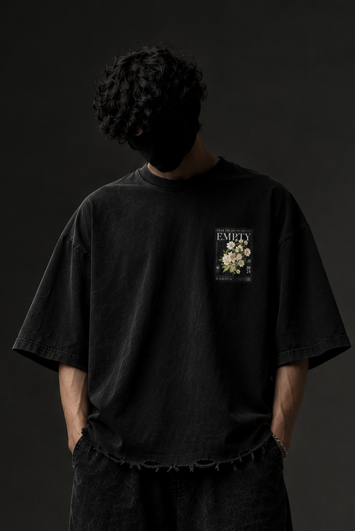 A person stands against a dark studio background wearing an oversized black graphic T-shirt, loose black pants, and black shoes. The shirt has a small chest graphic with the word 'VITALITY' and a floral illustration.