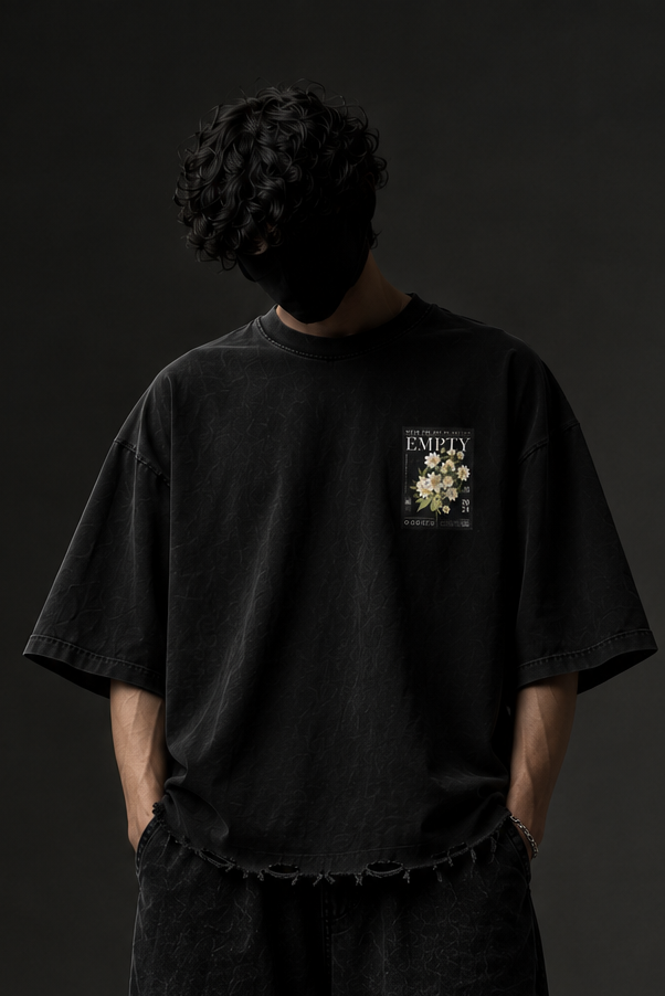 A person stands against a dark studio background wearing an oversized black graphic T-shirt, loose black pants, and black shoes. The shirt has a small chest graphic with the word 'VITALITY' and a floral illustration.